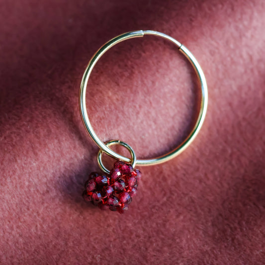A gold-colored jump ring with a red beaded heart puff charm attached to it, displayed against a red textured background.