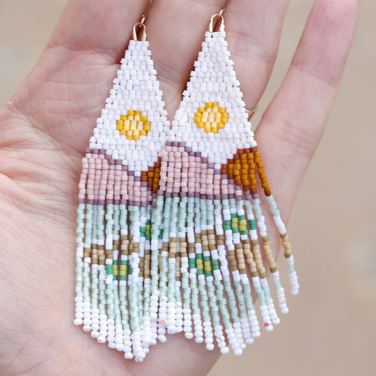 A pair of beaded earrings with gold filled findings, featuring white as the primary color with geometric patterns in yellow, green, and blue, held in a hand.