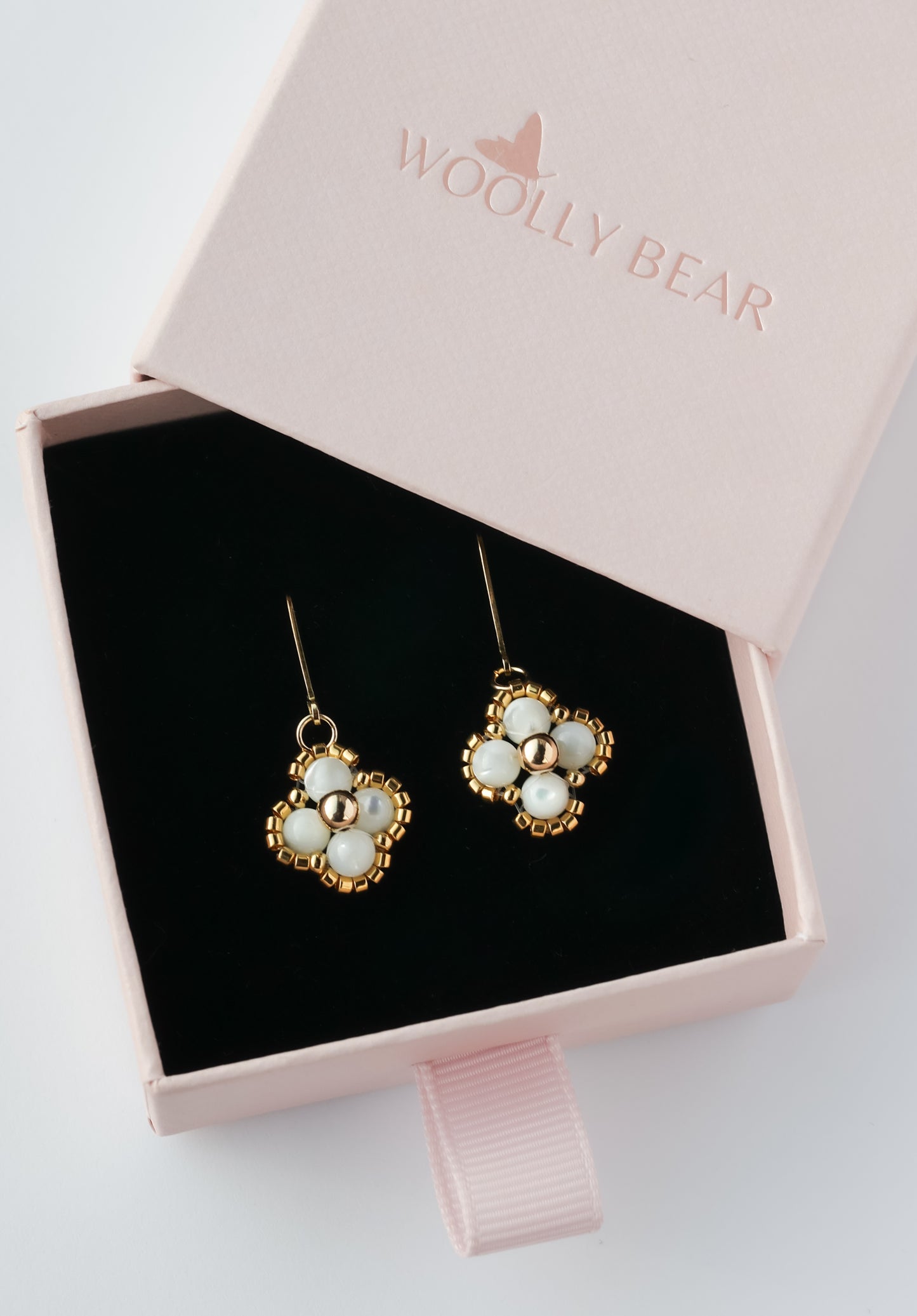 Gold and white floral beaded earrings in a pink 'Woolly Bear' box on a light gray background