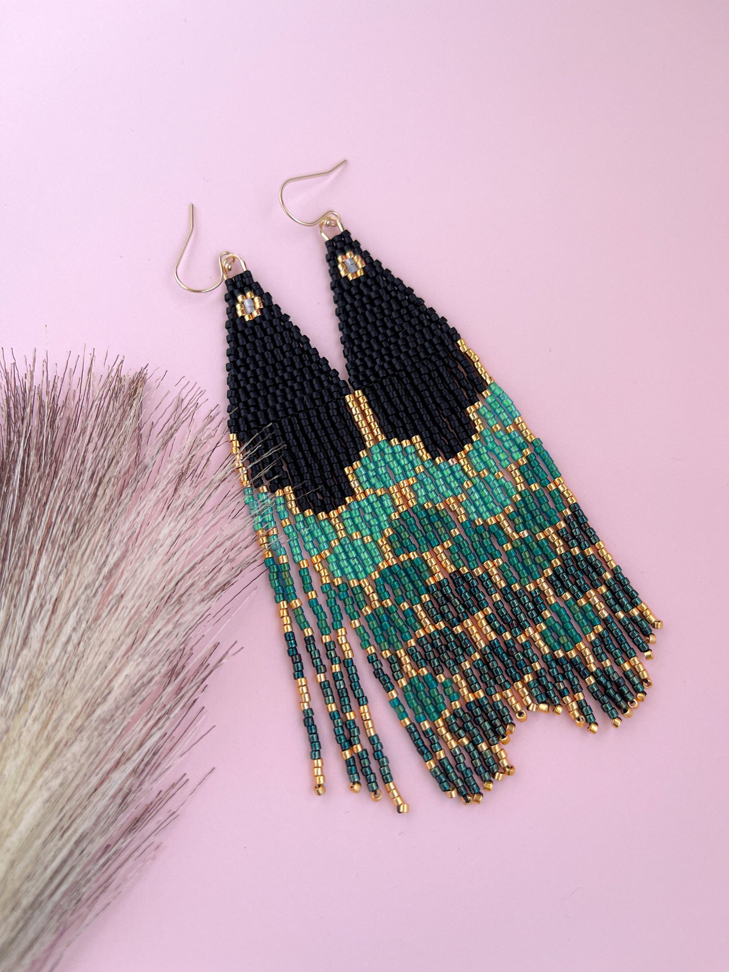 A pair of emerald green and black scalloped mermaid tail glass earrings with gold plated beads, displayed against a pink background with a decorative brush to the side.