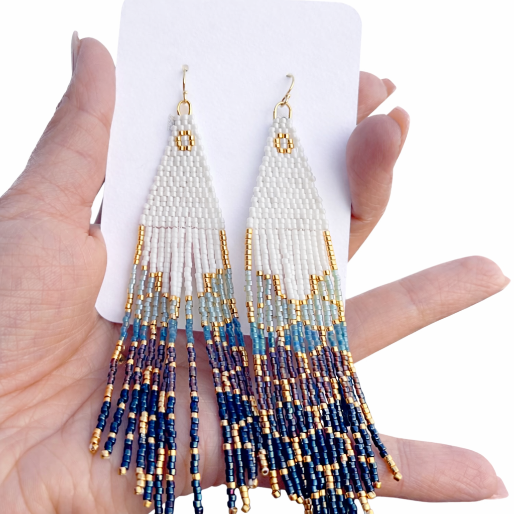 Handmade beaded earrings with white, blue, and gold colors held between fingers on a white background