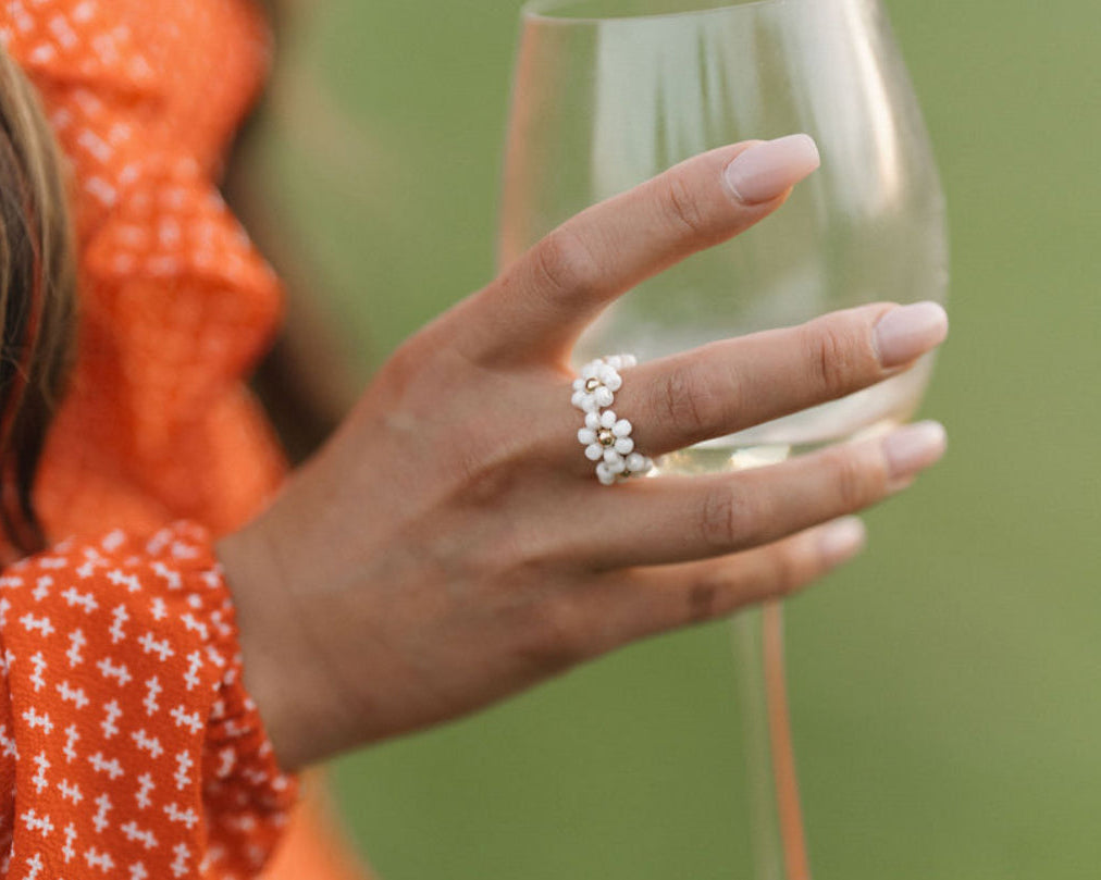 Person holding a glass with a blurred green background, wearing a handmade beaded ring