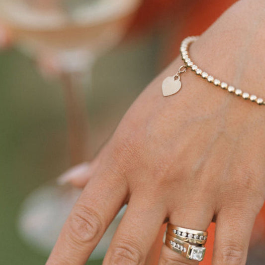 Hand wearing a gold bracelet with a heart charm and multiple rings against a blurred background