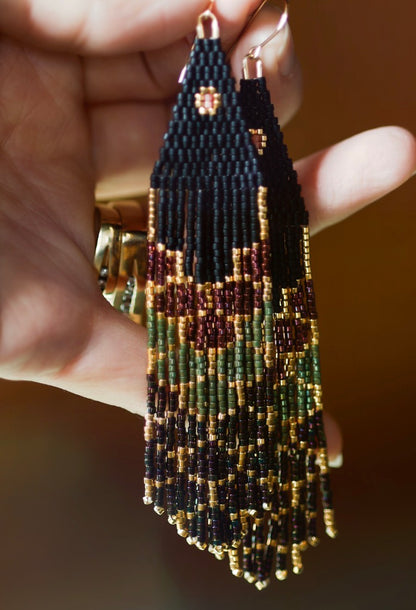 Hand holding handmade beaded earrings with black, gold, and green colors against a blurred background.